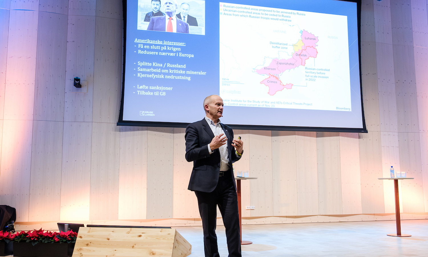 mann i mørk dress snakker mot publikum og gestikulerer med armene. I bakgrunnens nyst et lerret som viser grafer og tekst 