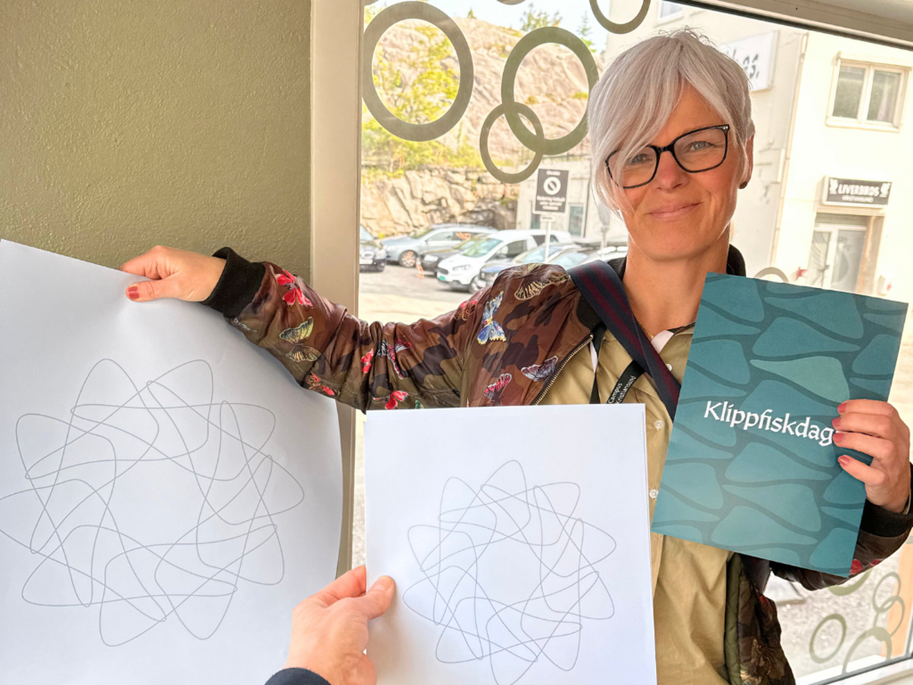 Bildet viser en kvinne med kortklippet, grått hår i en stilig frisyre, og med mørke briller som smiler mot kamere mens hun holder opp en plakate det står 