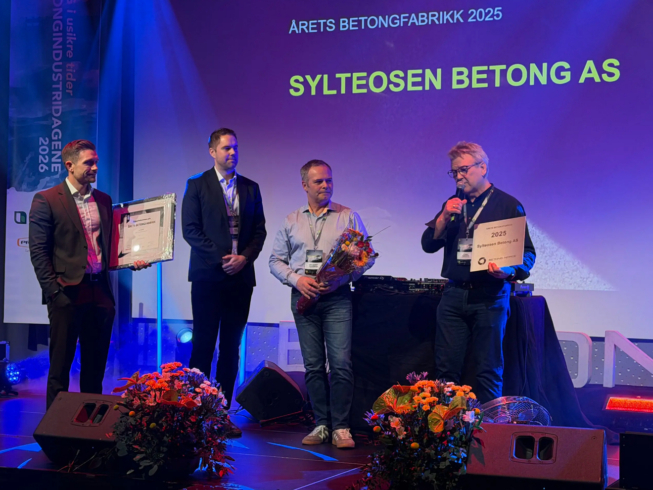 Fire menn står på en scene. Bak dem lyser en lilla skjerm opp. På den står det: at Sylteosen Betong AS har blitt kåret til Årets Betongfabrikk 2025