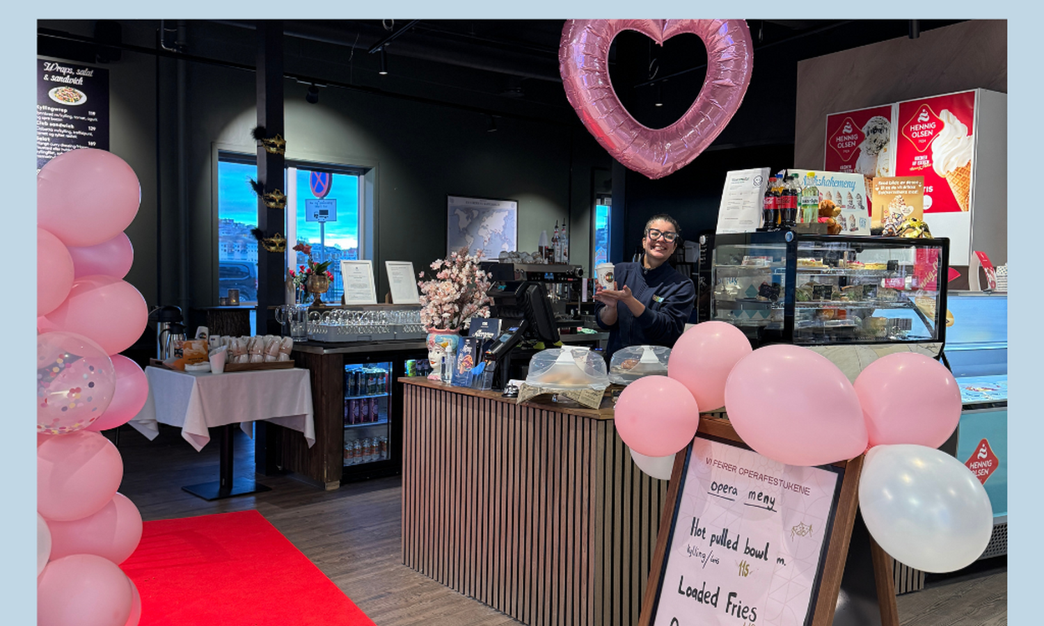 Kvinne bak en cafédisk omgitt av rosa ballonger, viser frem en brun pappkopp og smiler