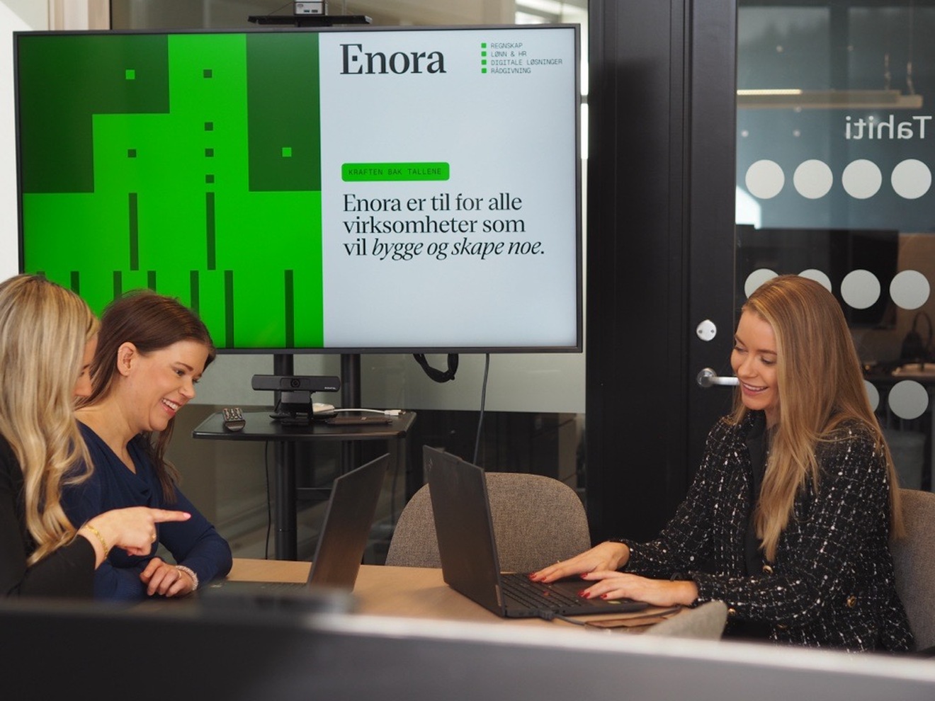 Tre kvinner sitter rundt et bord og ser inn i hver sin laptop  mens de smiler. 