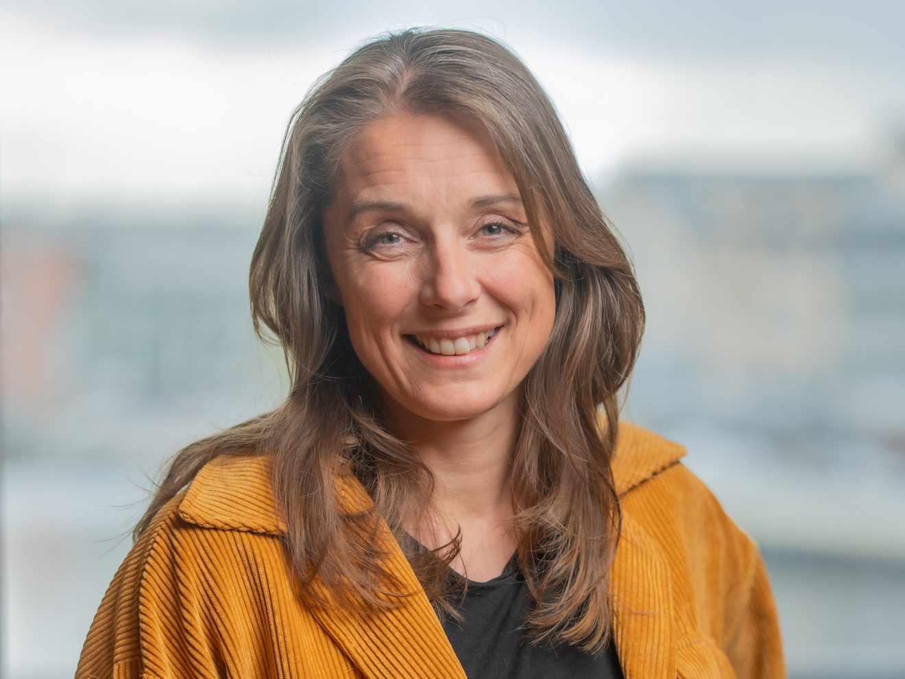 Kvinne med skulderlangt brunt og gråspettet hår smiler mot kamera. hun er ikledd en sennepsgul sjorte. 
