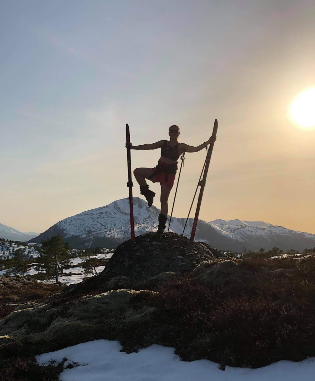 Kvinne står på toppen av en fjelltopp med avdre fjelltopper i bakgrunnen. Hun balaserer på en fot og støter seg med en ski i hver hånd